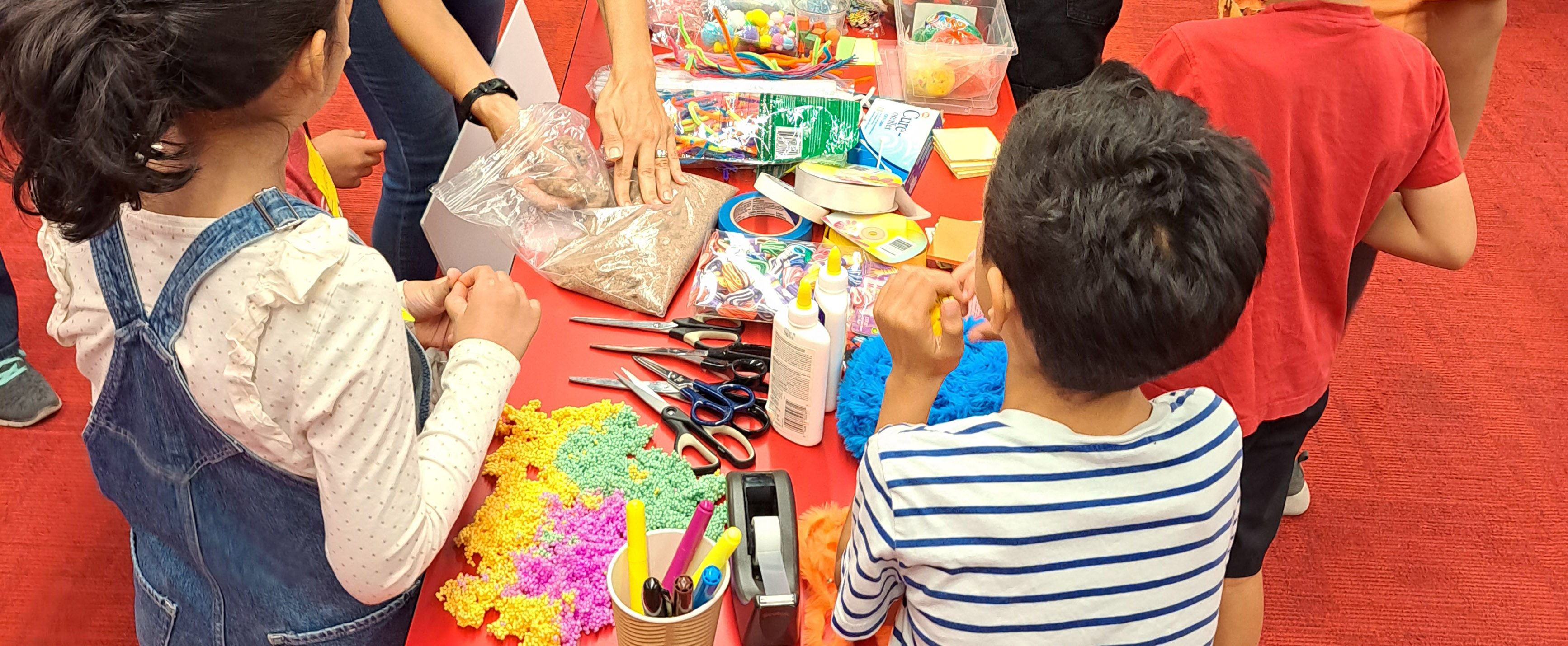Offereing a large selection of art and craft material to kids at once can be triggering for some children Art and craft tools and material on a red desk with kids standing around the table.